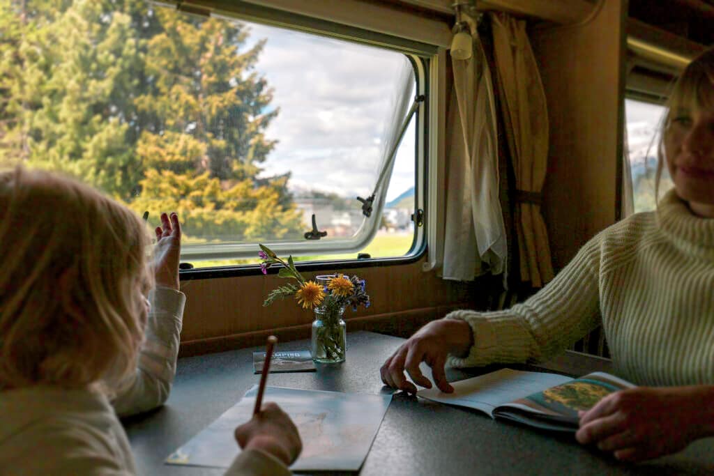 Familie im gemütlichen Mietwohnwagen auf dem Campingplatz St Moritz mit Blick auf die Natur durch das Fenster während ein Kind malt und eine erwachsene Person entspannt liest